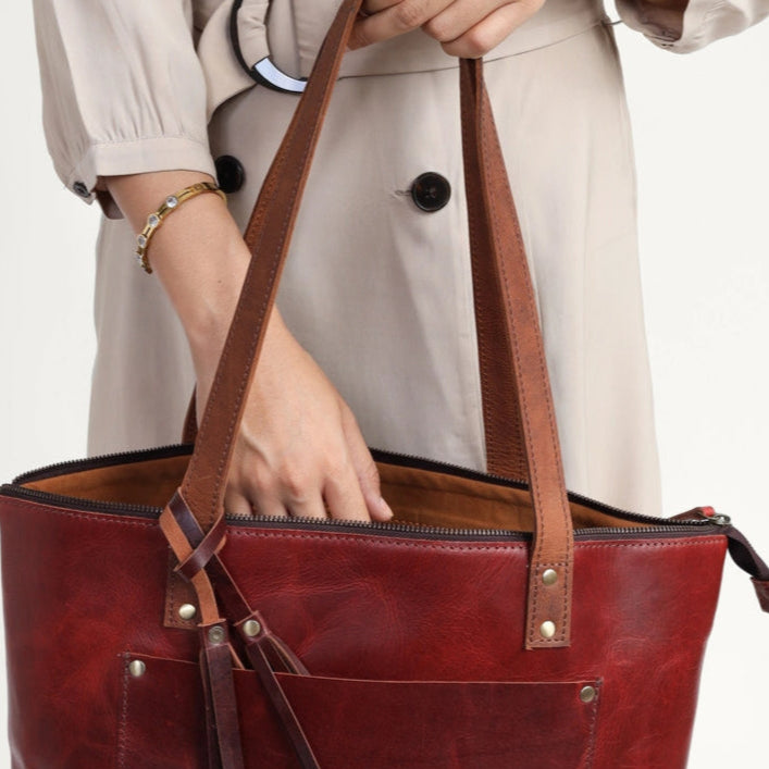wine leather tote bag held by a person wearing a beige trench coat on a light gray background